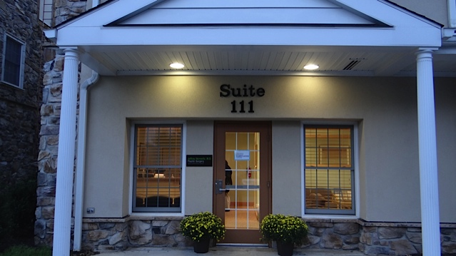 The image shows a building with an entrance sign labeled SUITE 111 above a doorway, featuring a covered porch with a white roof and columns, and the facade appears to be made of stone or brick.