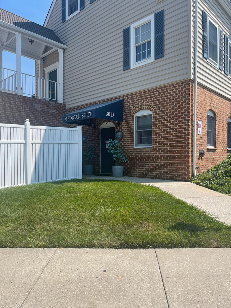 The image shows a two-story residential building with a brick facade, a white picket fence, and a green lawn. There is a sign on the building that reads AMERICAN SPRING, and below it, there s a smaller sign indicating a business or service within the building. The entrance has a black door and is sheltered by a small porch area with a bench. The front yard is neatly maintained with a lawn and a sidewalk leading to the entryway.