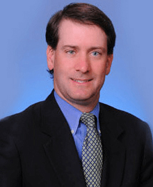 A man wearing a suit and tie, posing for a portrait with a blue background.