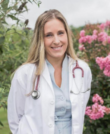 The image shows a woman wearing a white lab coat, standing outdoors with trees visible behind her, posing for a portrait.