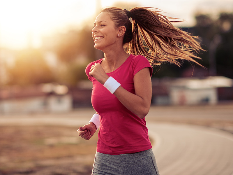 The image depicts a woman jogging with her hair flowing behind her, wearing a red tank top, black shorts, and a white visor. She appears happy and energetic, running on a dirt path during sunset.