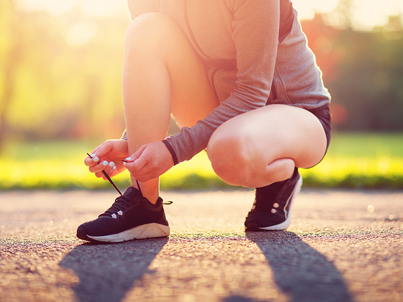 A person is kneeling on a paved pathway next to a shoe, adjusting its laces with their hands.