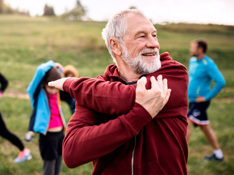 The image shows an older man wearing a red hoodie and white beard, stretching his arm behind him with a smile on his face, surrounded by several people outdoors during the daytime.